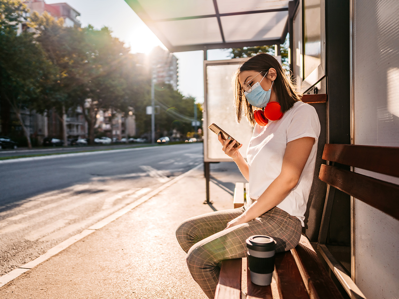 Woman with mask sitting at bus stop looking at phone