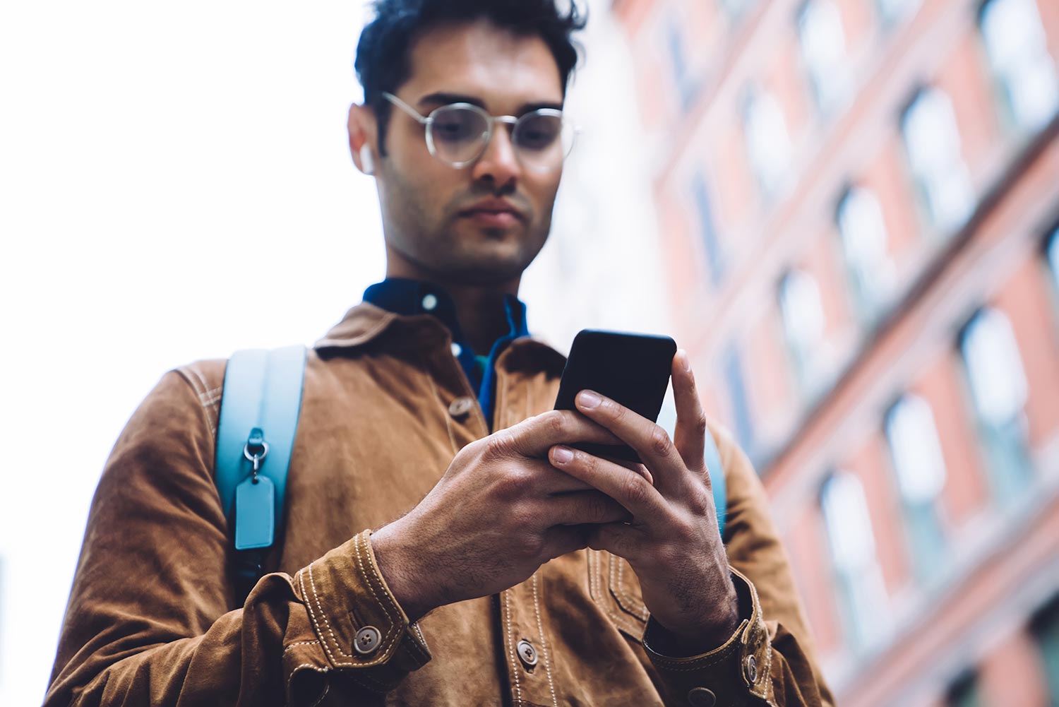 Indian man walking in the city with glasses looking down at his phone in his hands.