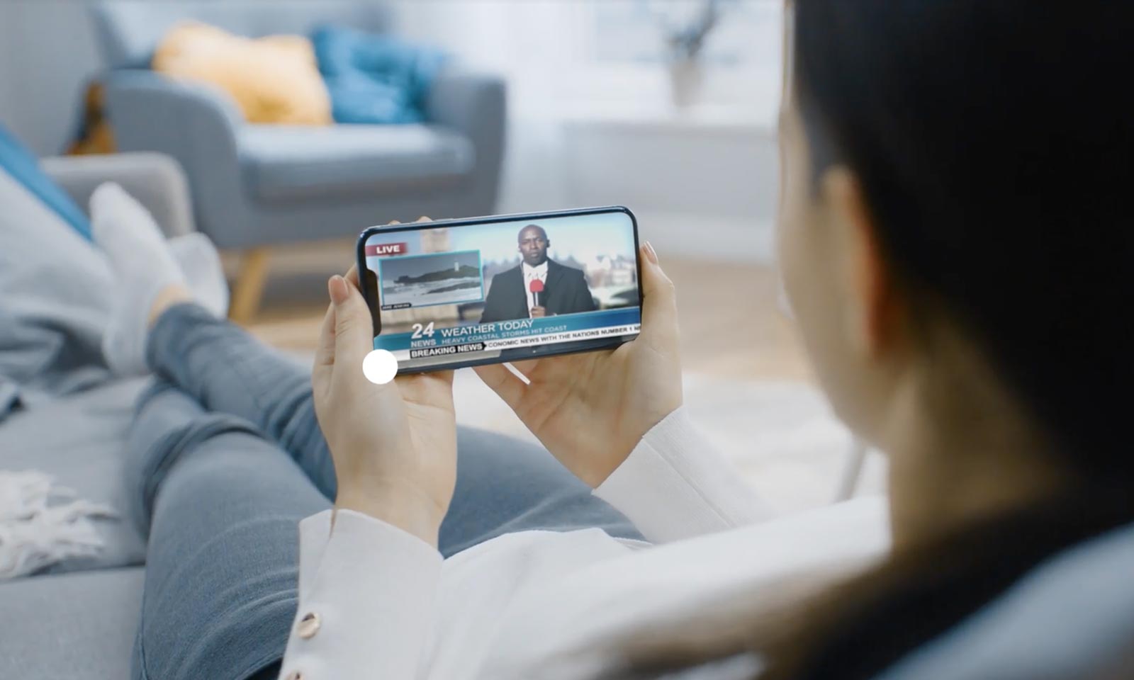 Woman lounging on couch watching a weather report on the phone in her hand.