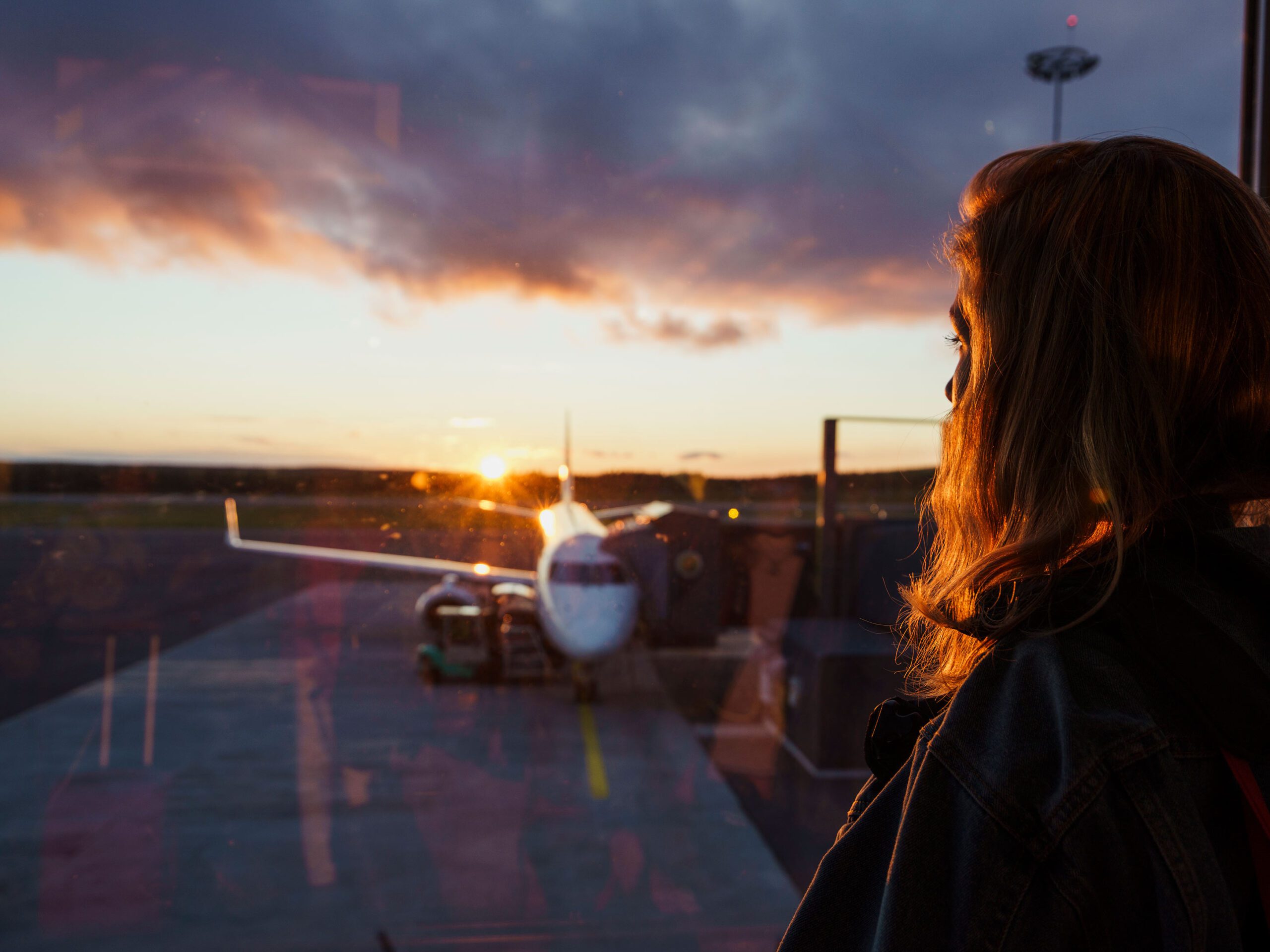 Young woman looking through window on plane at the airport at sunset