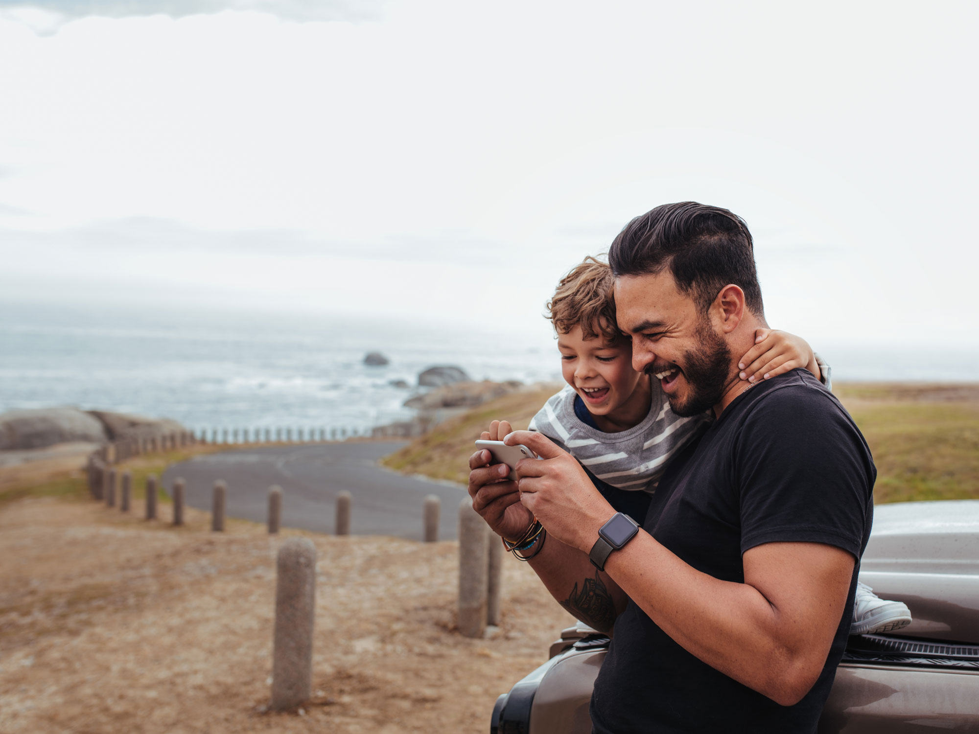 Father and son looking at phone and laughing while leaning against a car outside near a beach.