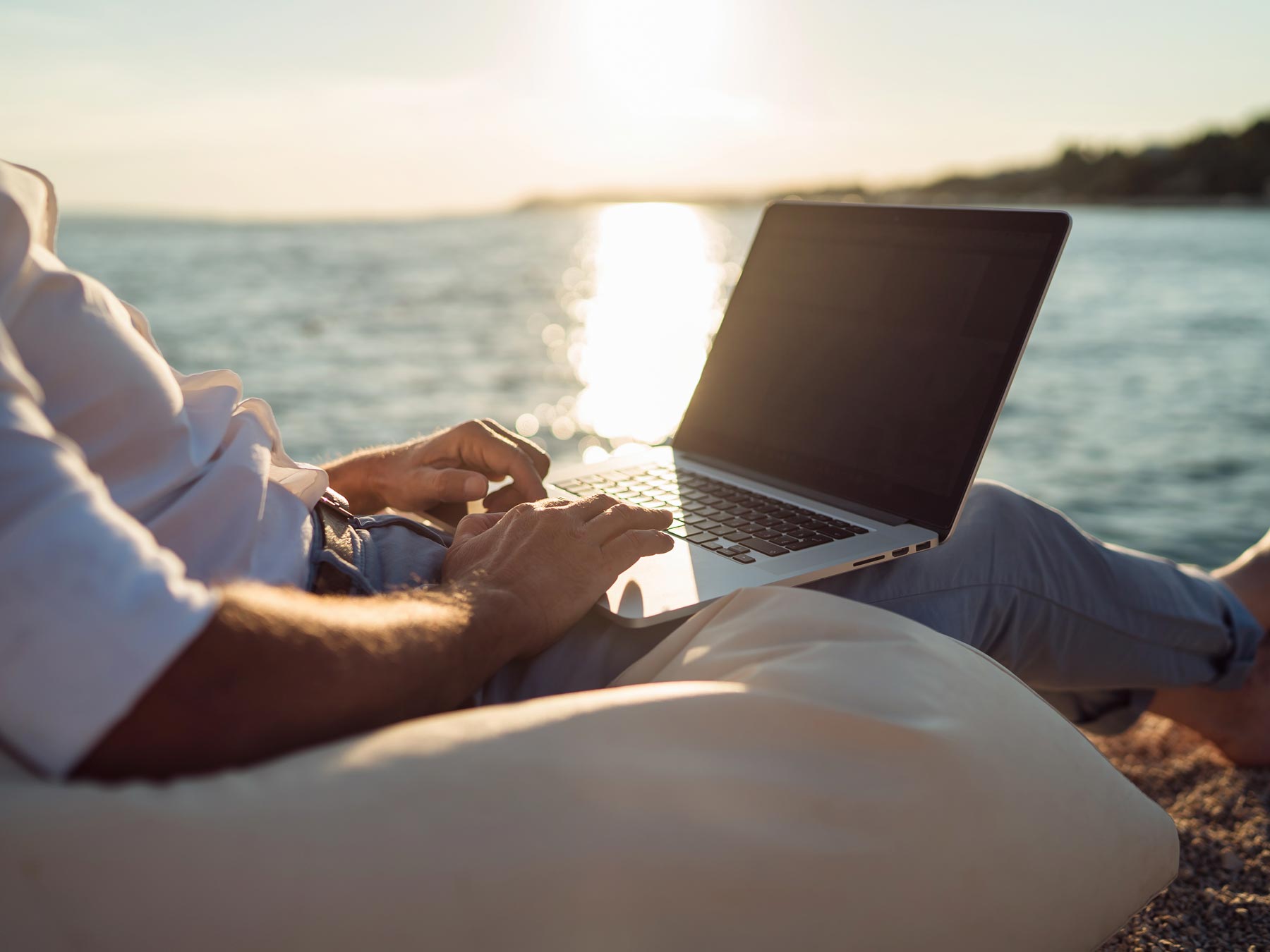 Man sitting on beach a beach chair with laptop in his lap and the sun shining behind him.