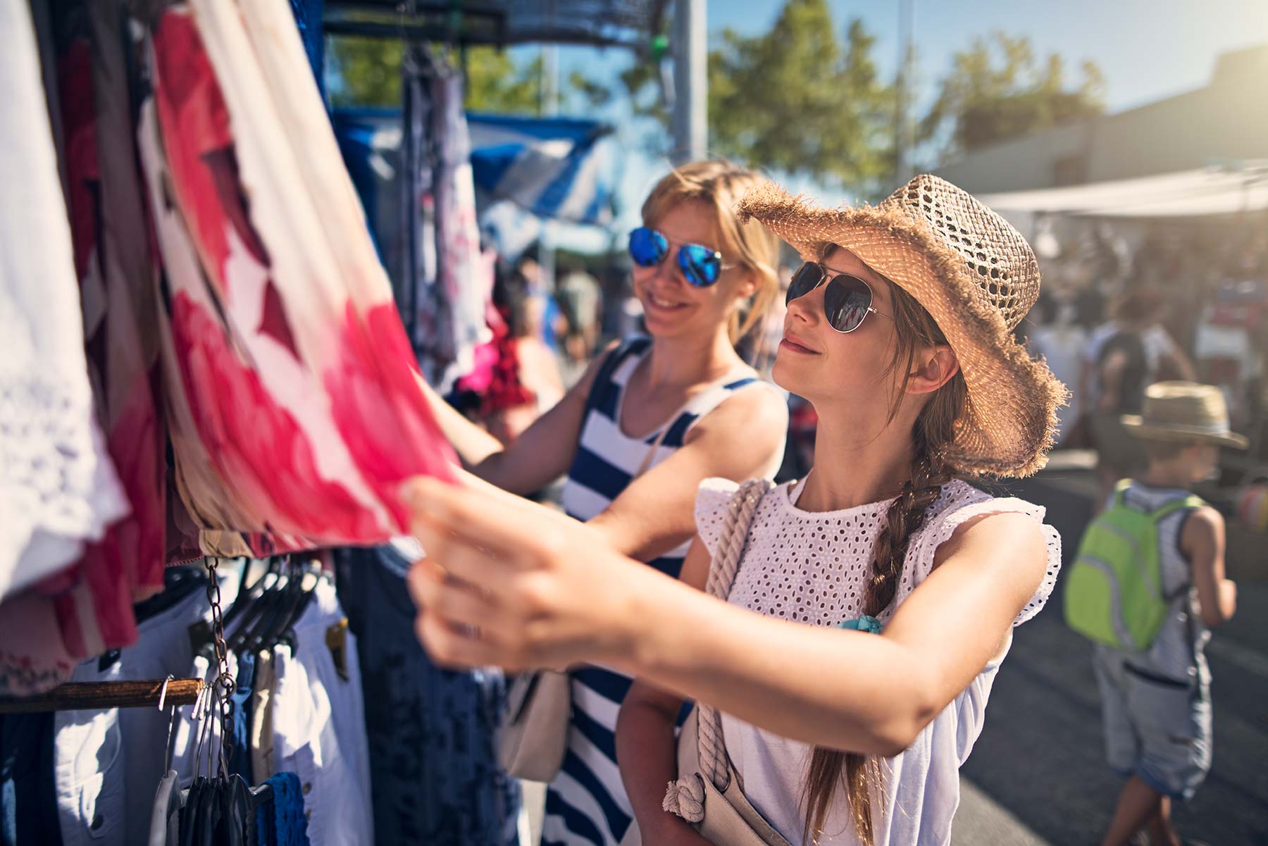 Two women looking at clothing on a rack outdoors on a sunny day sunglasses and a hat on.