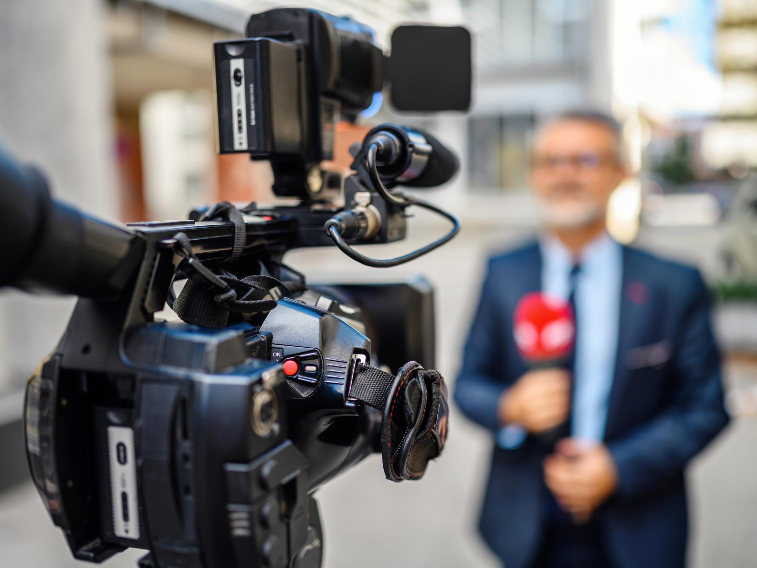 A male television reporter holding a microphone, is standing outdoors in front of a building. A male camera operator is filming him