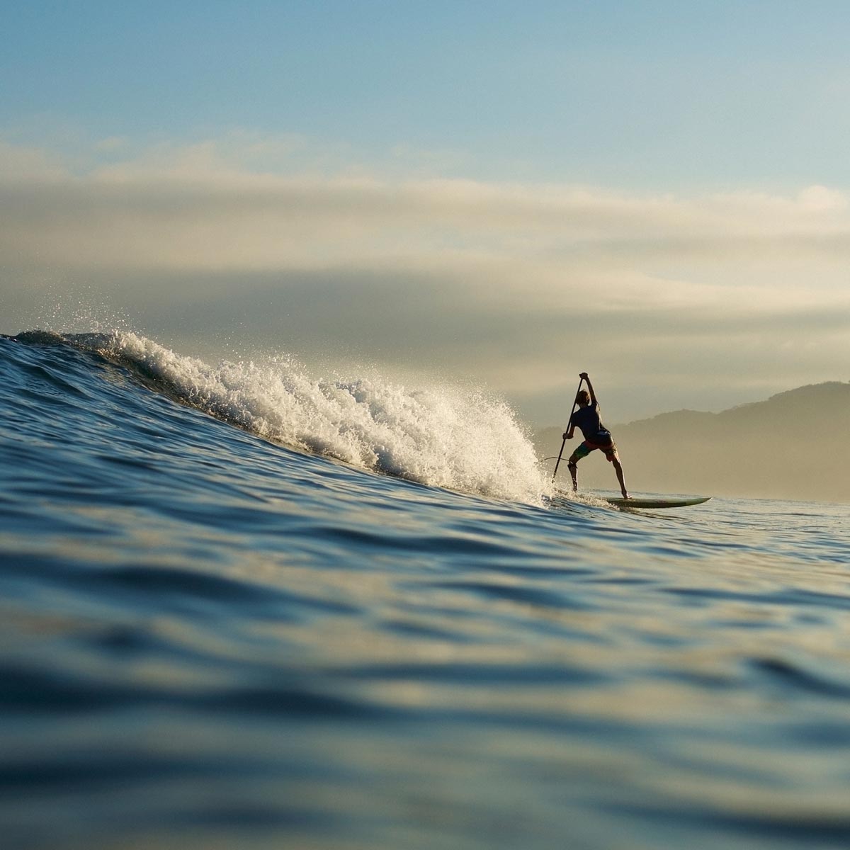 Surfer seen in the distance riding a wave with beautiful clouds and hills in the background.