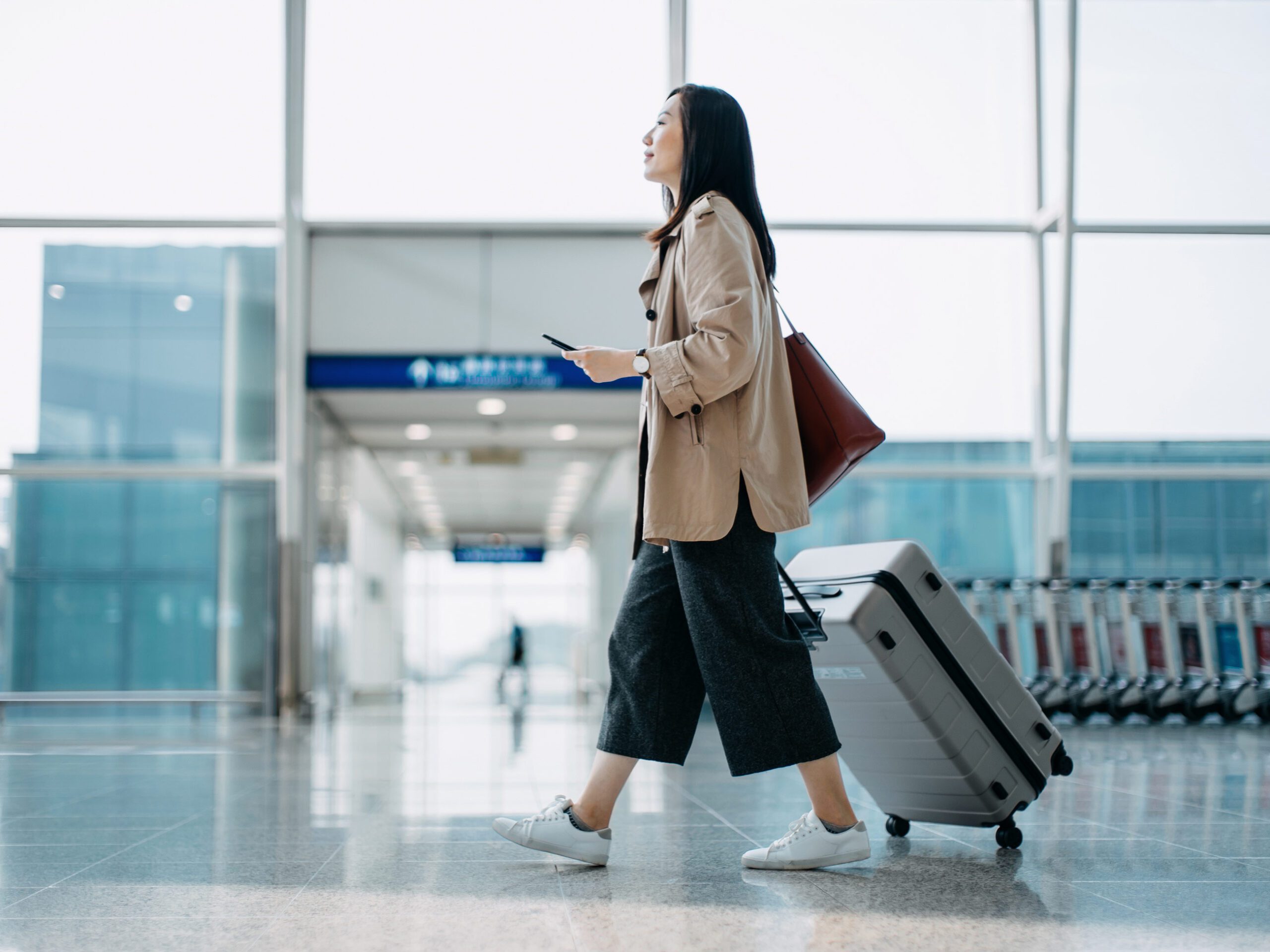 Young Asian woman carrying suitcase and holding smartphone, walking in airport terminal