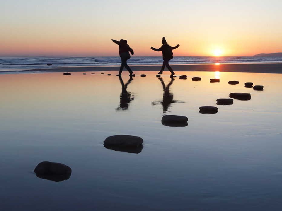 A couple on beach jumping across stones during sunset