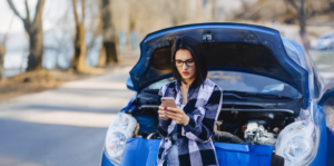 Woman on her phone standing in front of her car that has its hood open.
