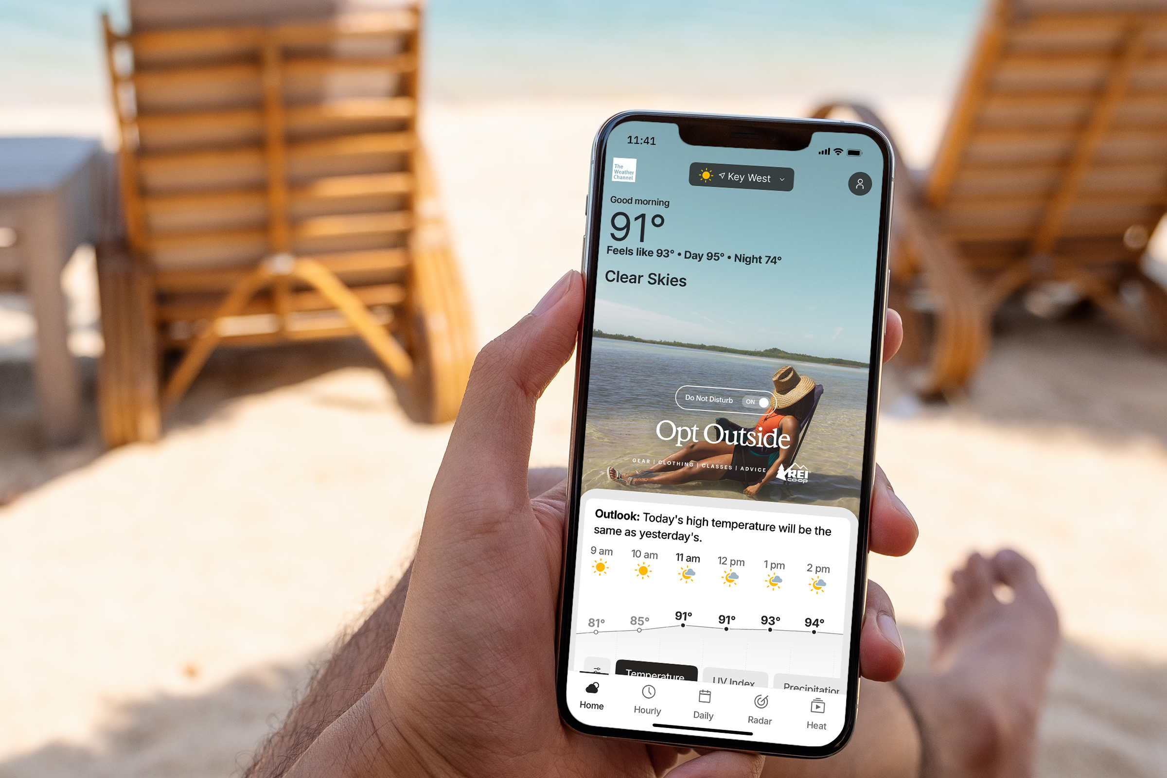 A person at the beach on a sunny day looking at The Weather Channel app on their mobile phone which shows a sunny day REI advertisement