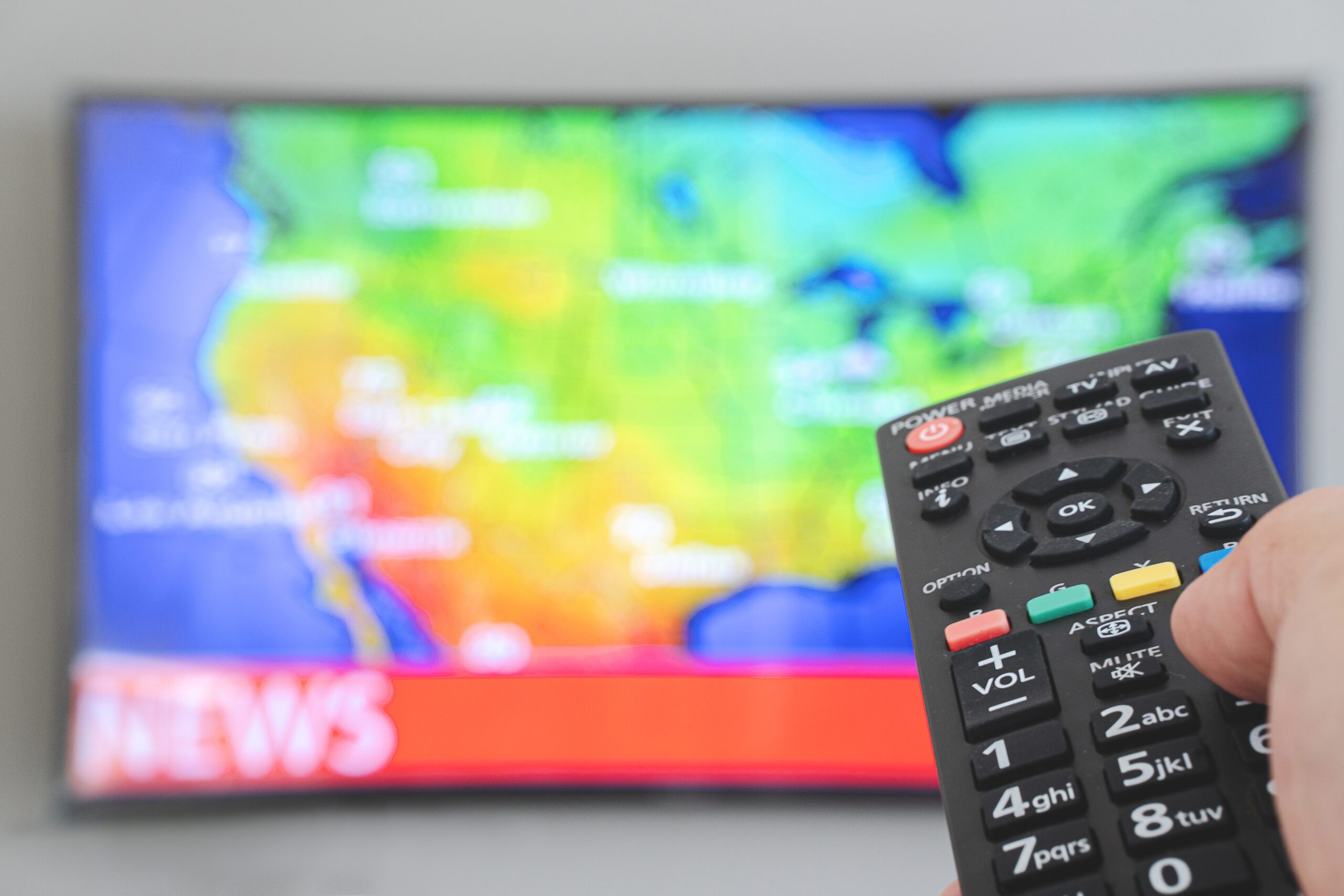 Hand holding TV remote control with a television weather map in the background.