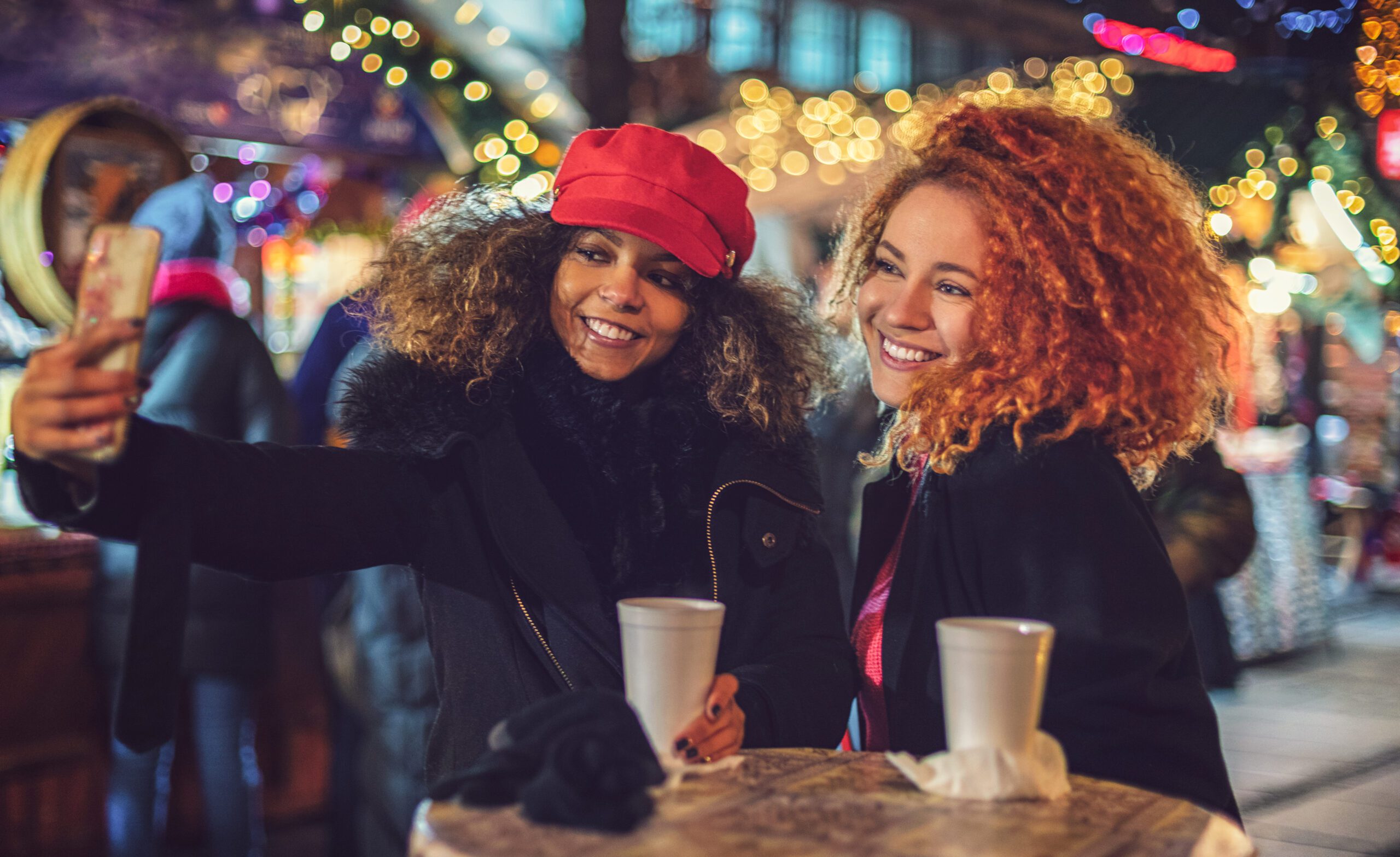 Two Gen Z women are shopping with hot drinks at a holiday market, taking a selfie photo.