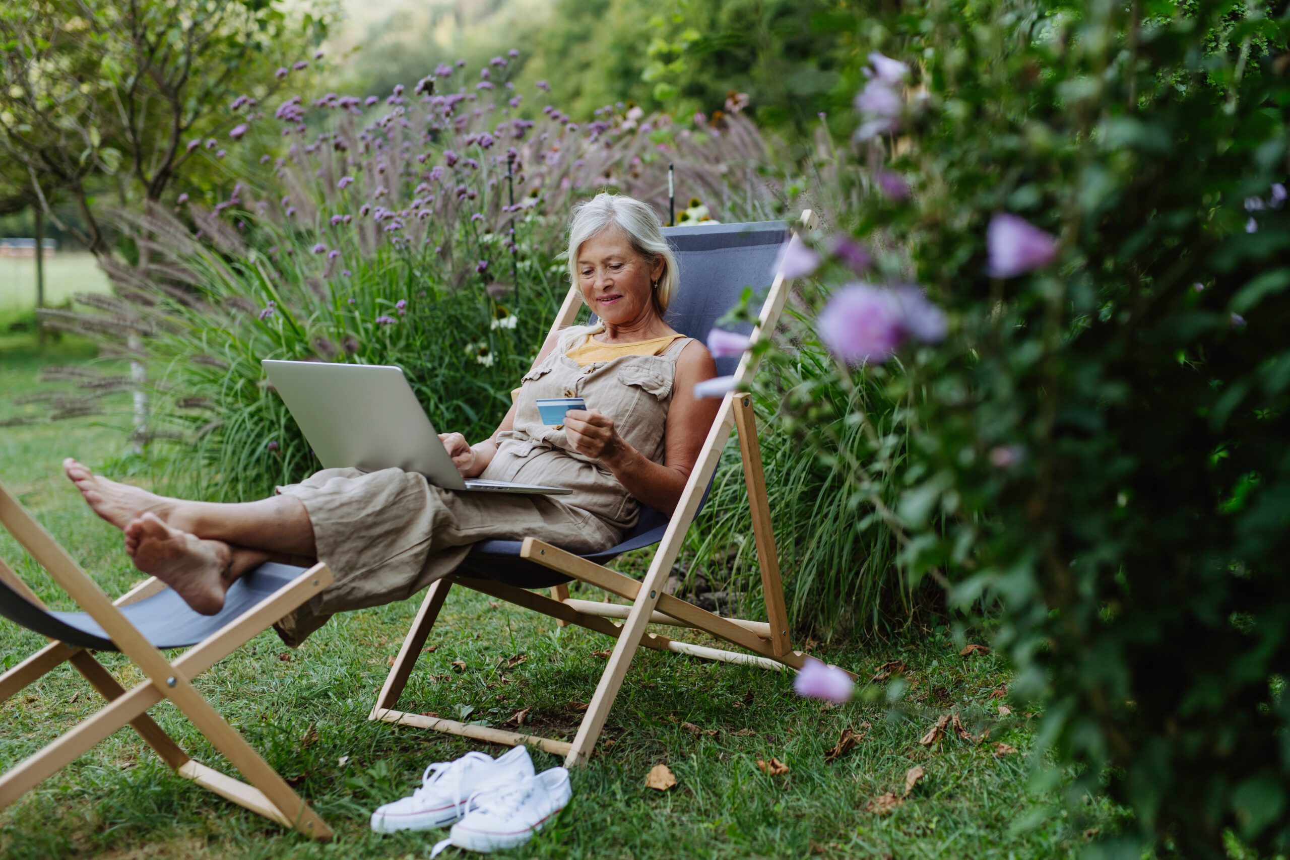 Older woman sitting outdoor on deck chair, shopping online on notebook. Woman holding debit card in hand, imputing card detail for payment.