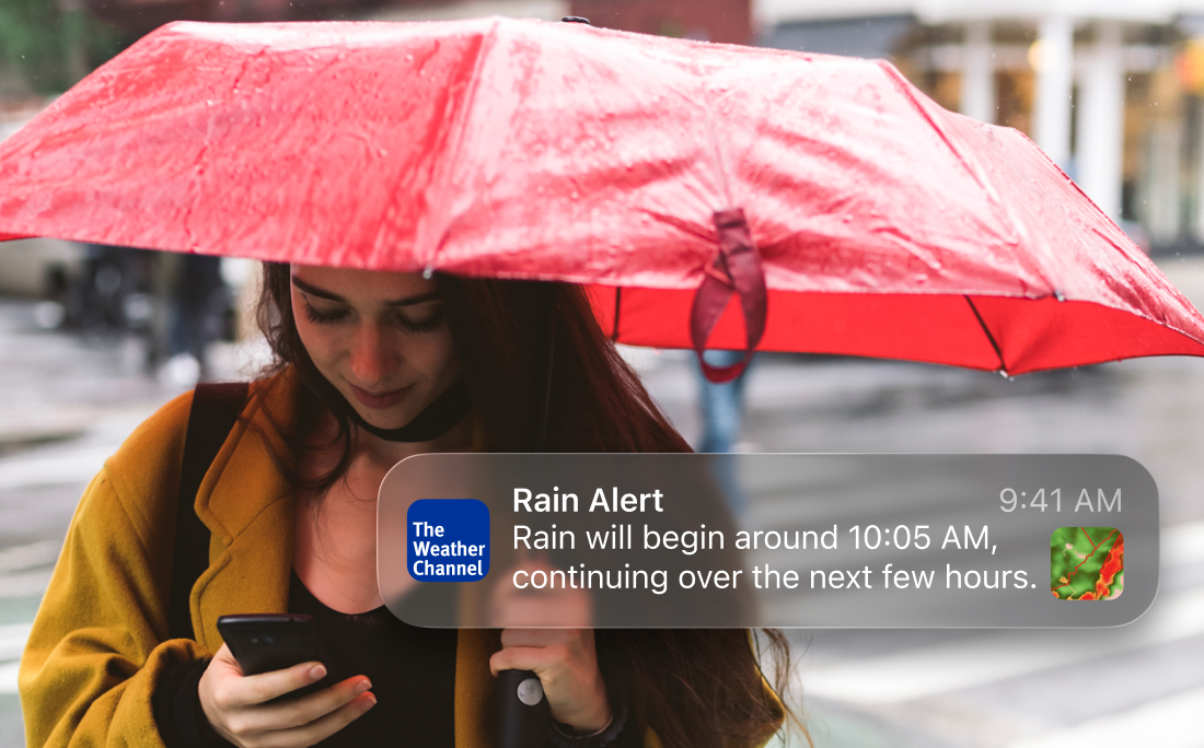 A woman under an umbrella in the rain reading a weather alert on her mobile phone.