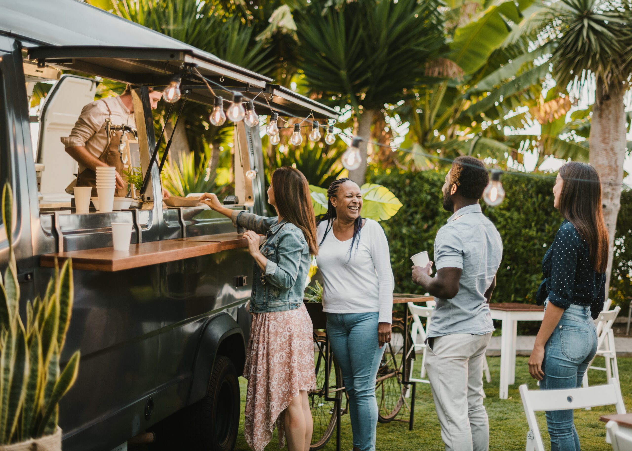 Happy woman smiling and chatting with diverse friends while standing in queue near food truck in park on summer day