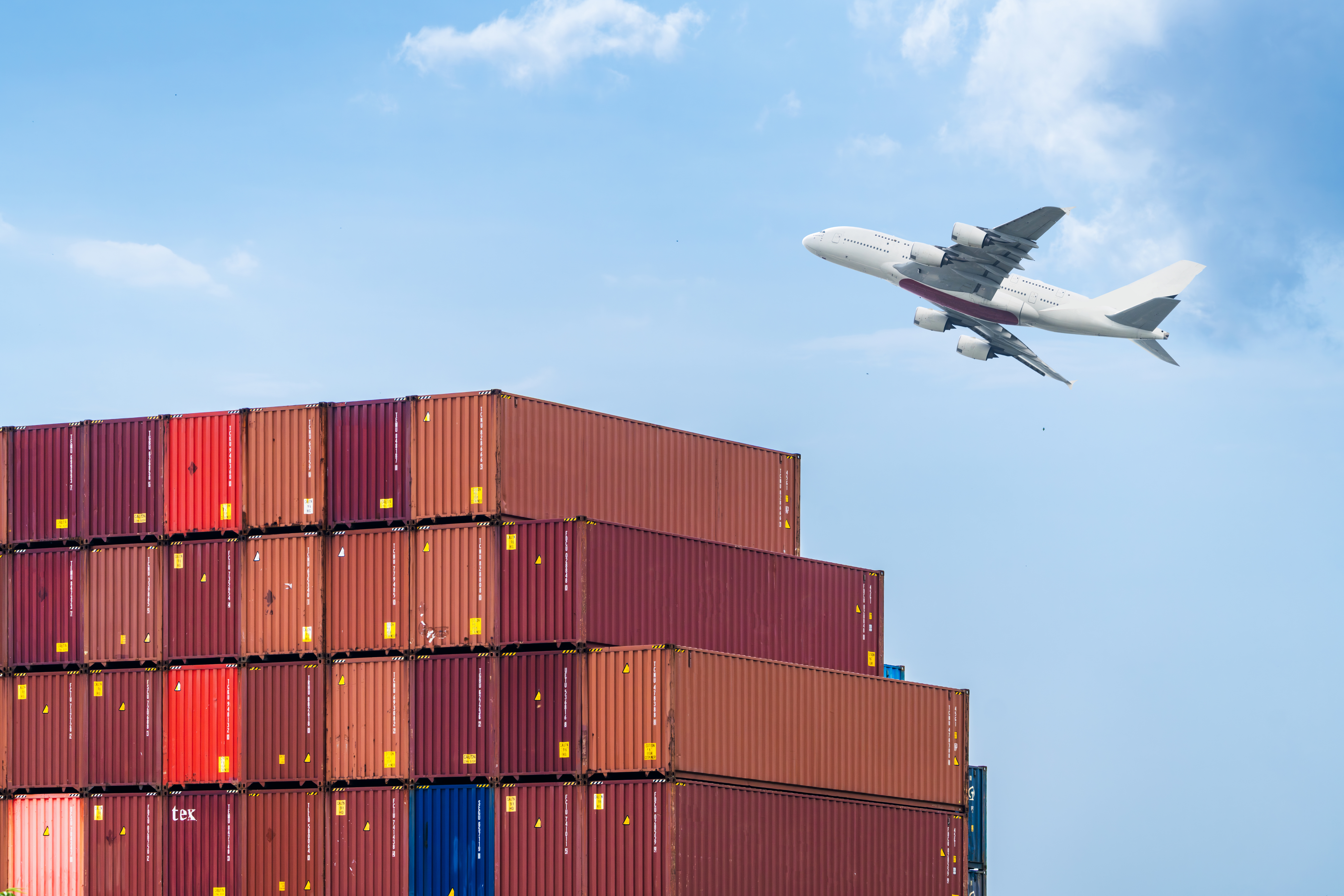 A commercial airplane soars over colorful cargo containers stacked high, symbolizing international trade and logistics. The clear sky enhances the theme of seamless global transportation.